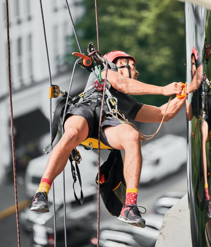 Industrial mountaineering worker cleaning window outside building.